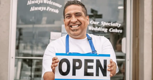 Small business owner holding an 'Open for Business' sign with a confident smile, symbolizing successful management and efficient scheduling through tools like RosterElf.